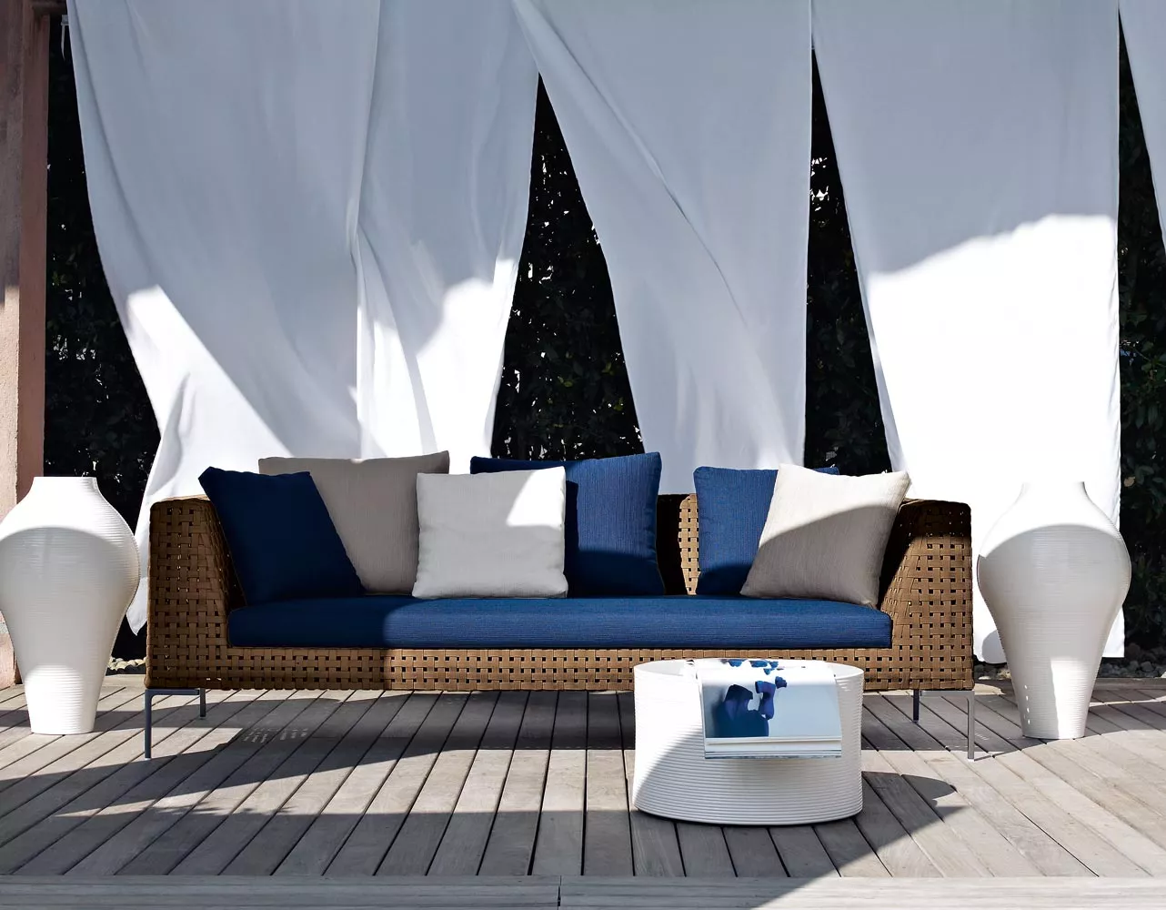 Charles Outdoor Sofa von B&B Italia mit blauen und weißen Kissen auf einer Terrasse zwischen weißen Vorhängen und zwei weißen Vasen 