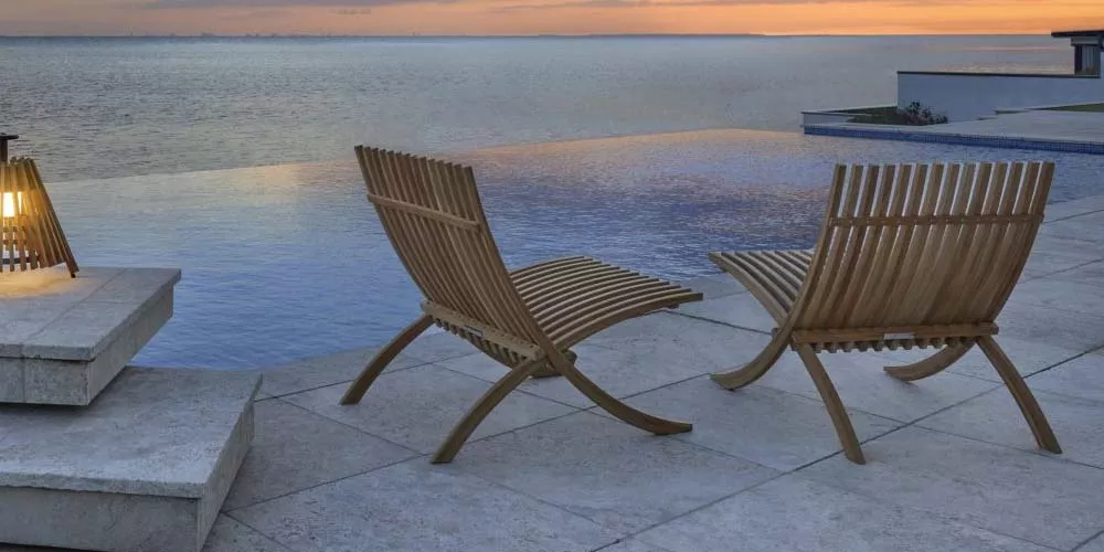 Zwei Skargaarden Nozib Stühle aus Holz auf einer Terrasse mit Blick auf einen Infinity-Pool und das Meer bei Sonnenuntergang.