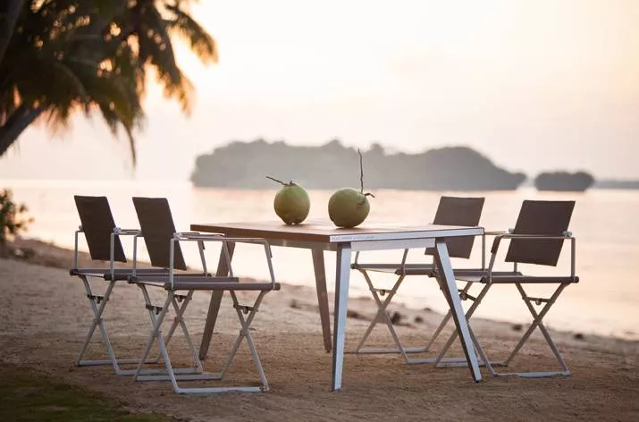 Dedon Seax Kollektion Seax Armlehnstuhl klappbar am Strand bei Sonnenuntergang mit Tisch und Kokosnüssen im Hintergrund Meer und Palmen