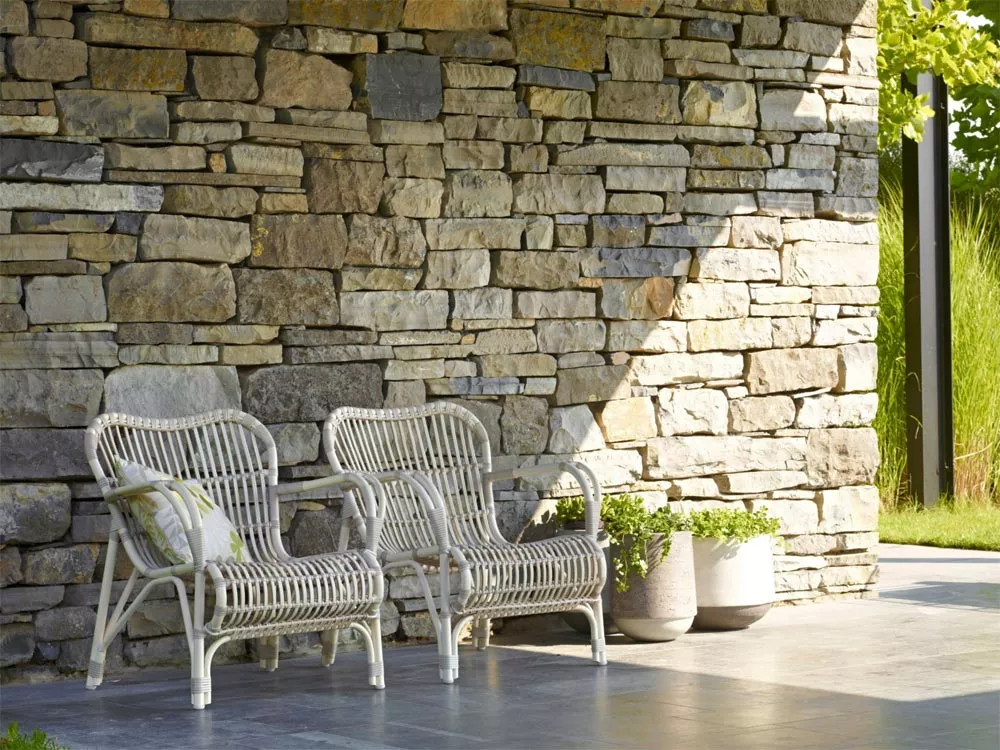 Zwei Vincent's Garden Lucy Gartensessel in Beige vor einer Steinmauer im Garten, mit Kissen und Pflanzen im Hintergrund.