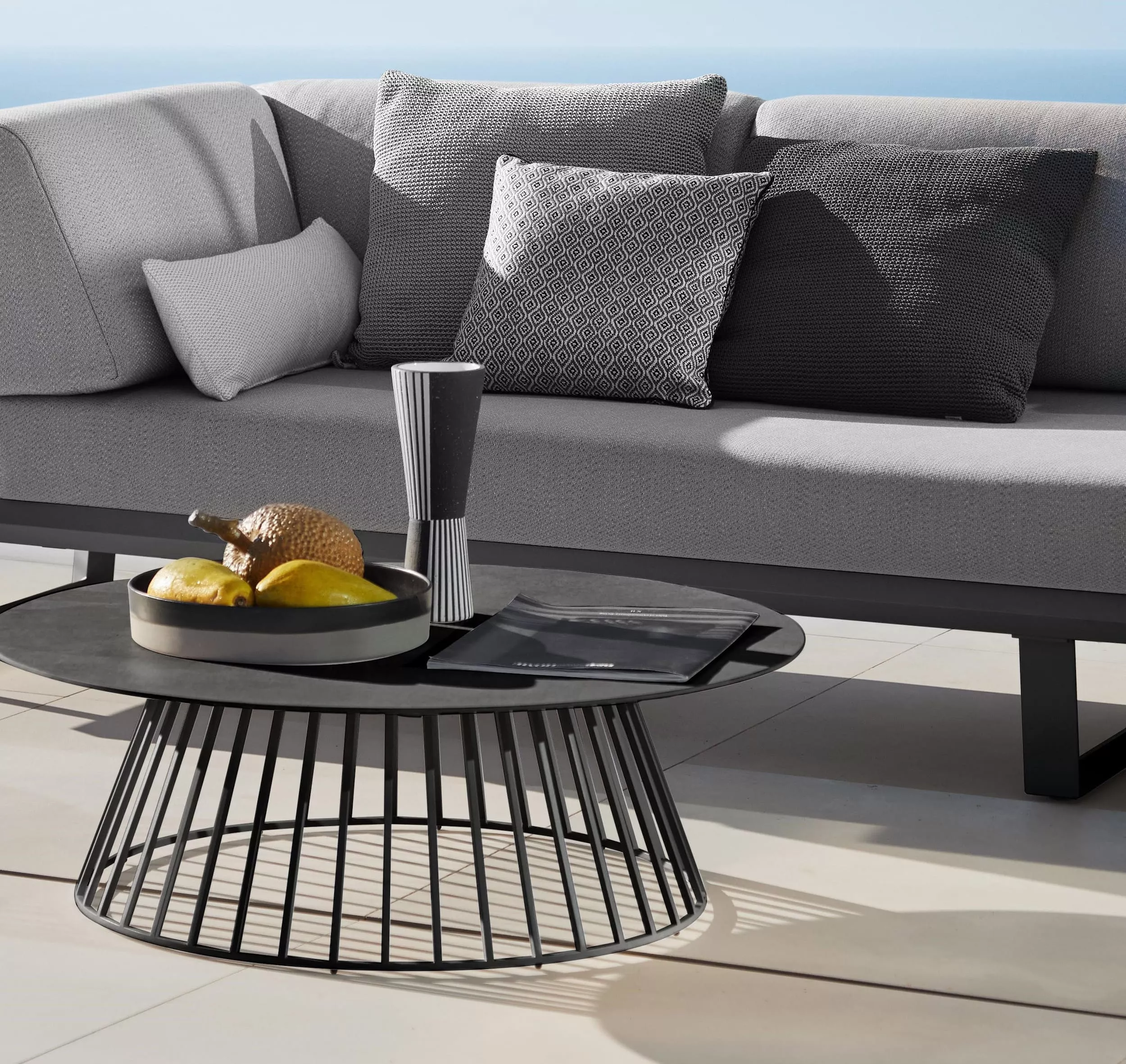Solpuri Boom Sofa mit grauen Kissen und einem modernen Couchtisch mit Obstschale und Vase in einer stilvollen Outdoor-Umgebung.