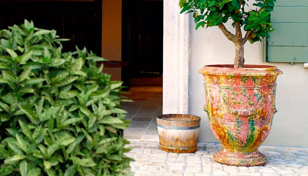 Großer, bunter French Pottery Topf im mediterranen Garten auf Kopfsteinpflaster, neben einem kleineren Holzeimer, vor einem Haus.