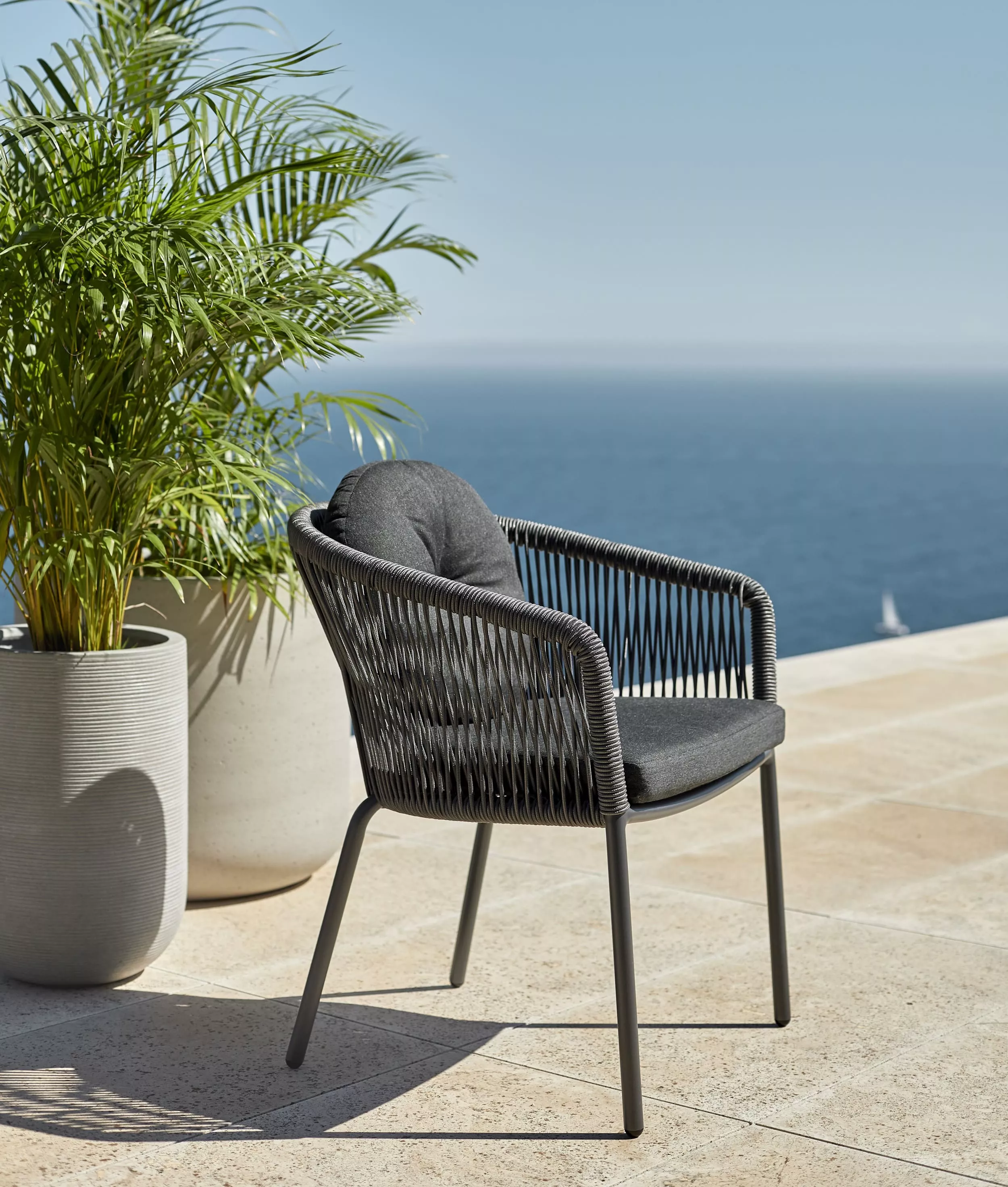 Ein Solpuri Loop Stuhl steht auf einer Terrasse mit Blick auf das Meer neben zwei großen Pflanzen in Töpfen im Hintergrund.