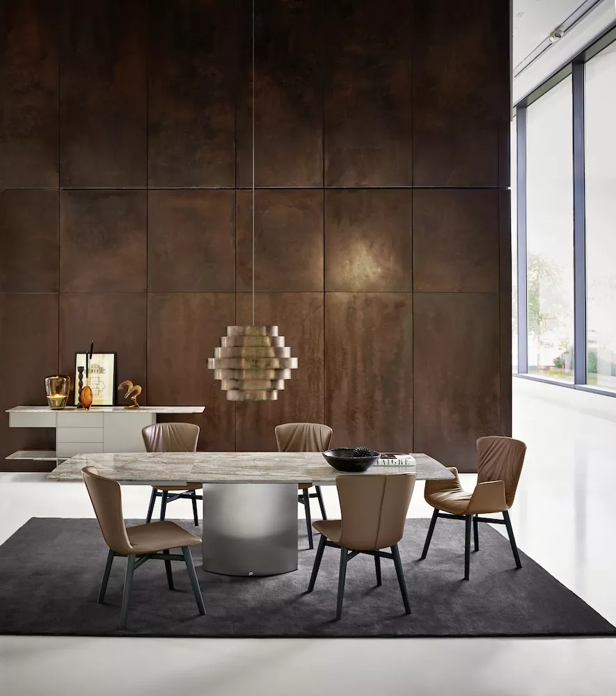 Brauner DRAENERT ADLER Tisch aus Stein in modernem Esszimmer mit großen Fenstern und stilvoller Beleuchtung auf dunklem Teppich.