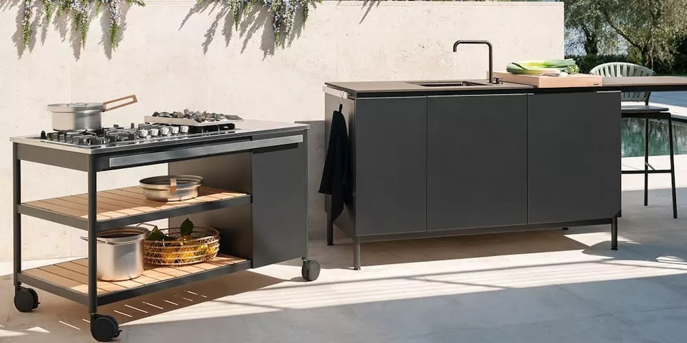 Außenküche von Villa Schmidt mit grauem Schrank und mobilem Grillwagen auf heller Terrasse, umgeben von Pflanzen.