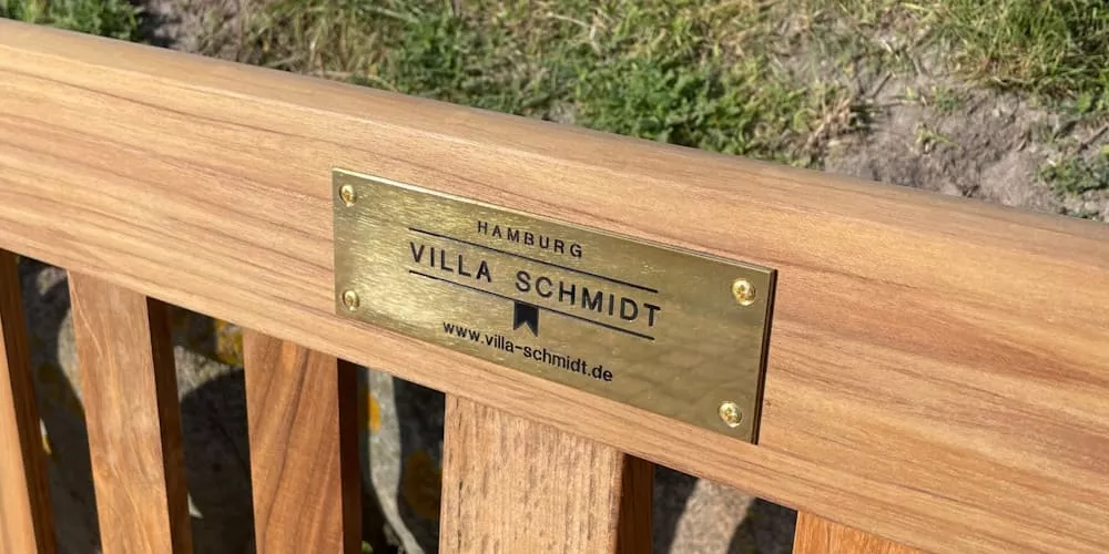 Holzbank mit Messingschild der Marke Villa Schmidt auf grünem Rasen im Sonnenlicht.