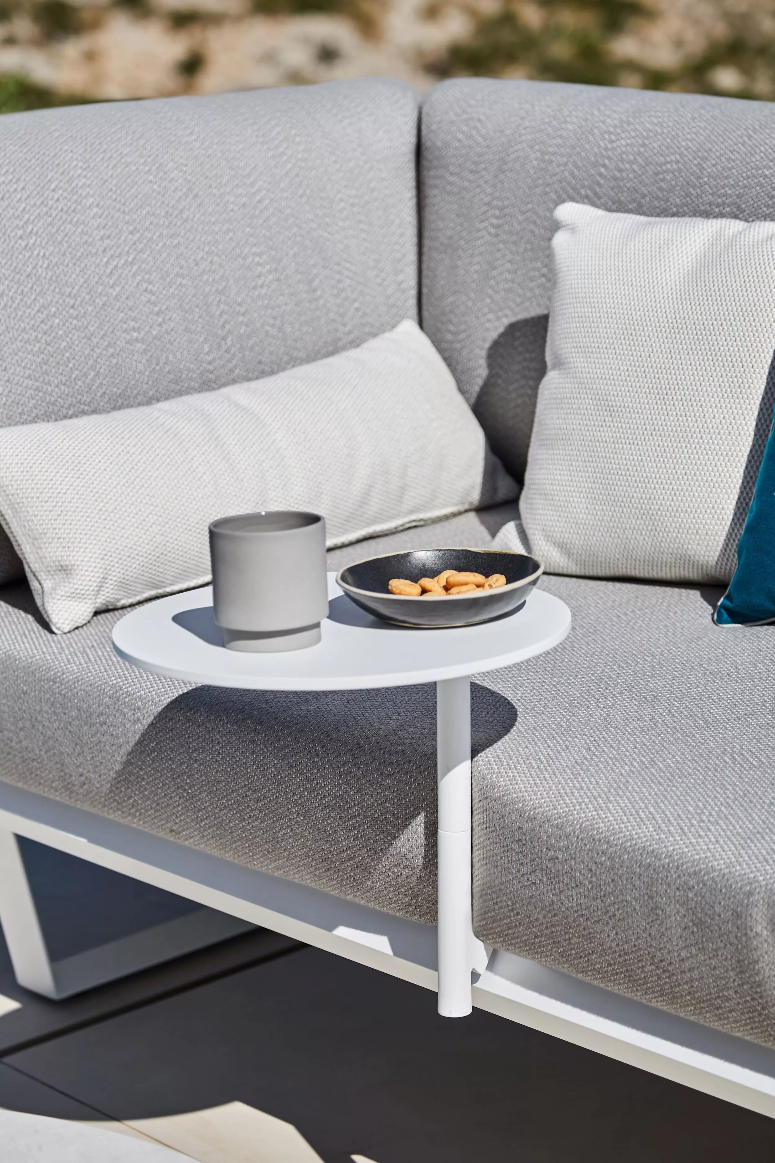 Solpuri Boom Sofa mit grauen Kissen und einem kleinen weißen Beistelltisch auf dem ein graues Gefäß und eine Schale mit Snacks stehen Moodbild im Freien