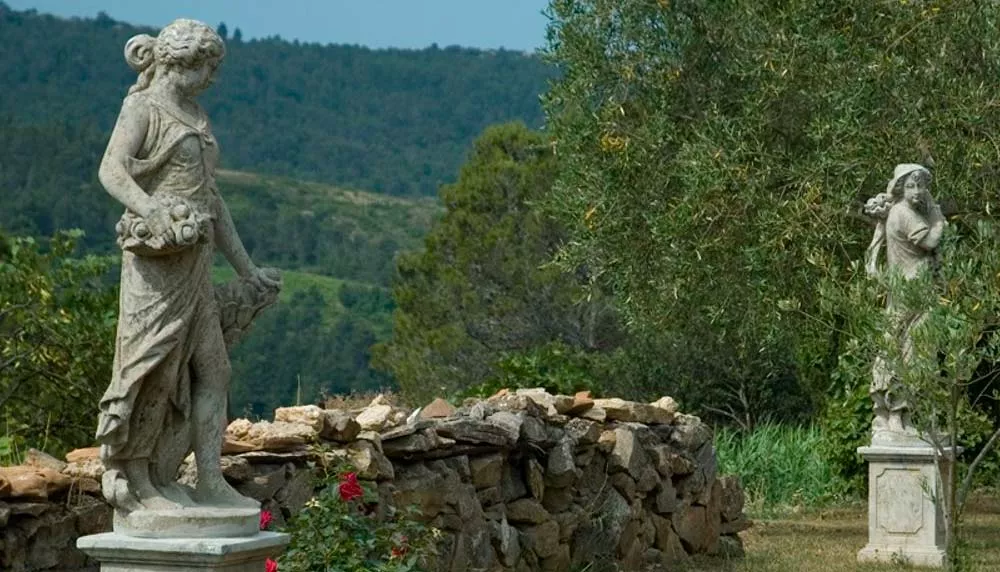 Zwei französische Steinstatuen in einem Garten vor einer malerischen Waldkulisse, umgeben von Natur und Blumen.
