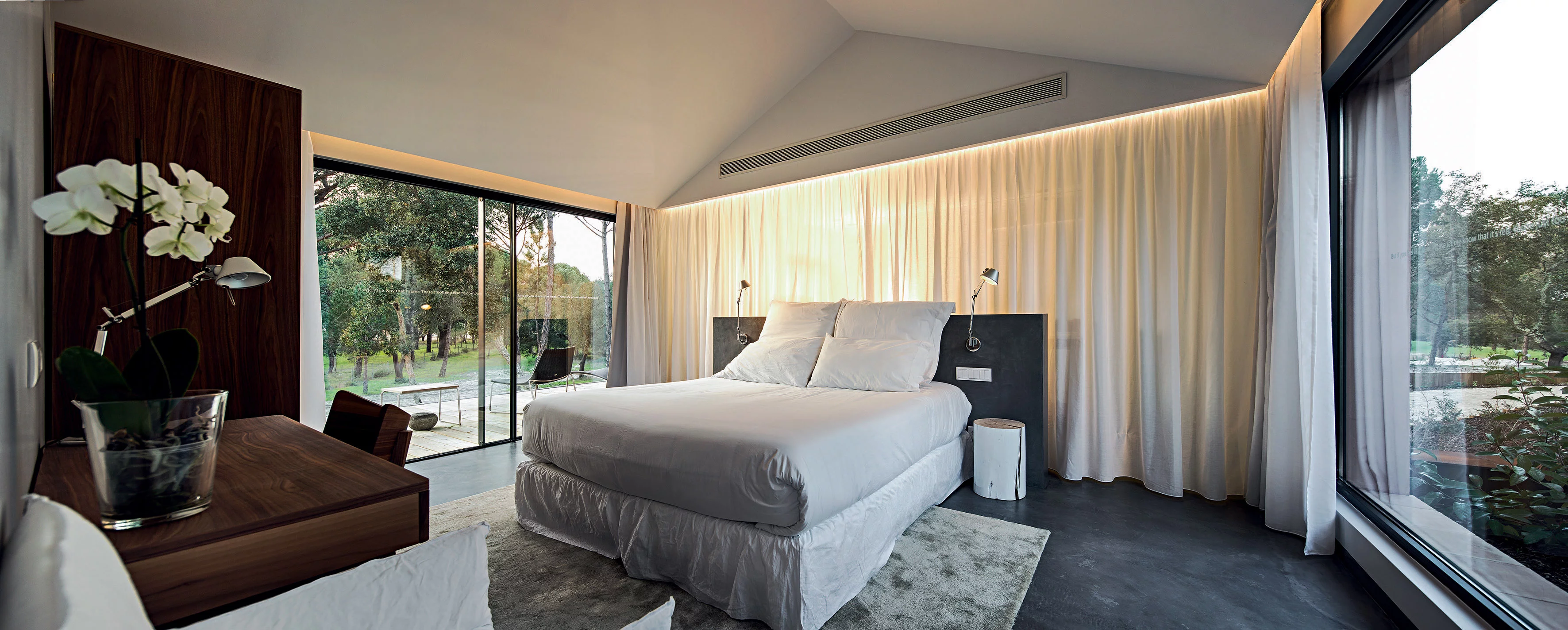 Ein modernes Schlafzimmer mit einem großen Bett und weißen Vorhängen mit Blick auf einen Garten durch große Fenster 