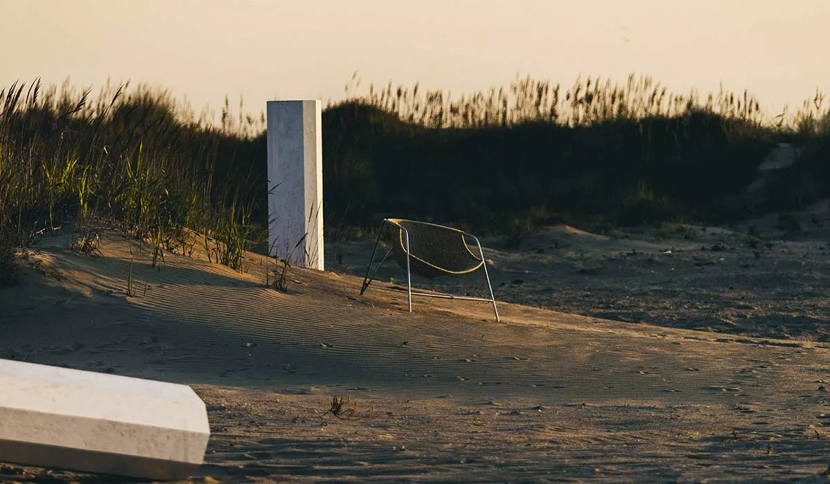 Calma Loungesessel in Beige auf sandigem Strand bei Sonnenuntergang, umgeben von Gräsern und Betonblöcken, stimmungsvolle Atmosphäre.