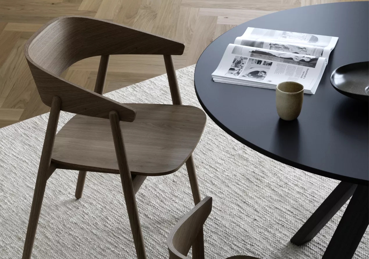 Stuhl aus der AC Kollektion von Villa Schmidt in dunklem Holz neben einem runden, schwarzen Tisch auf einem hellen Teppich.