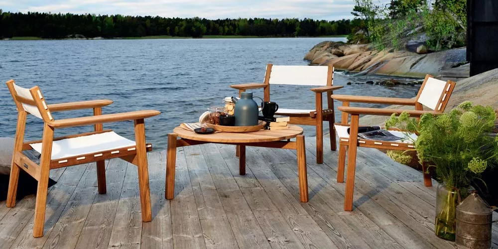 Skargaarden Djurö Outdoor-Möbelset aus Teakholz mit weißen Textilene-Sitzflächen auf einer Terrasse am See bei bewölktem Himmel.