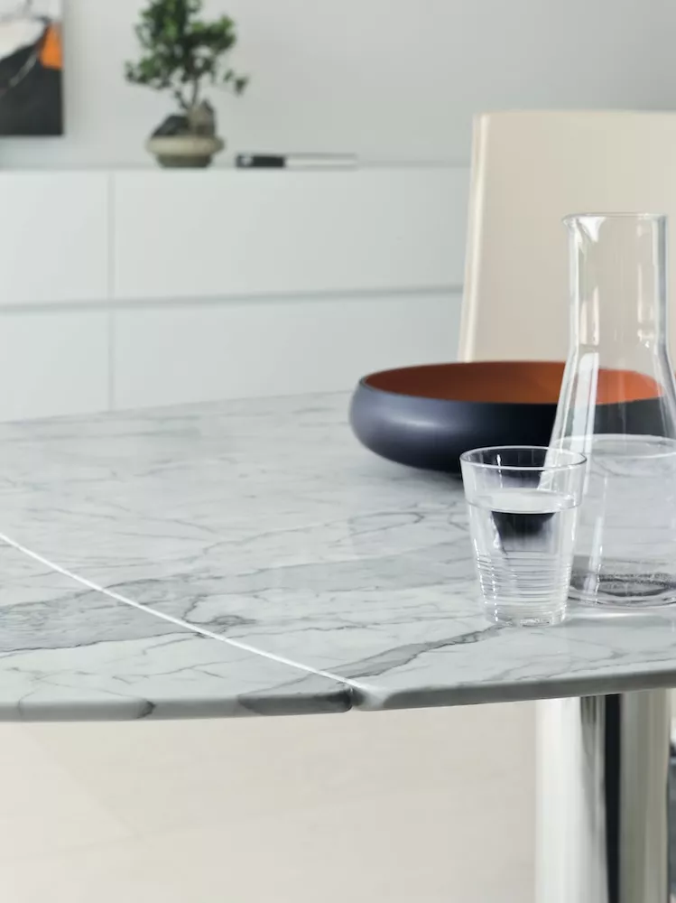 Ein Titan Draenert Tisch mit einer Steinplatte und einem Glas Wasser sowie einer Karaffe auf einem modernen Esstisch in einem hellen Raum.