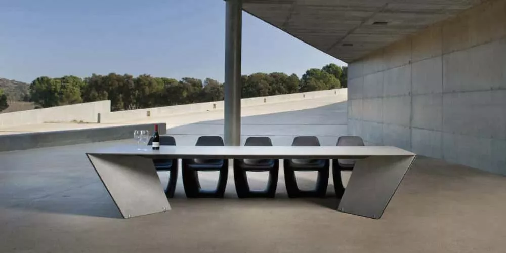 Calma Tisch, Modell Angle, in Grau, auf einer Terrasse mit modernem Betonhintergrund, mit sechs passenden Stühlen und einer Weinflasche.