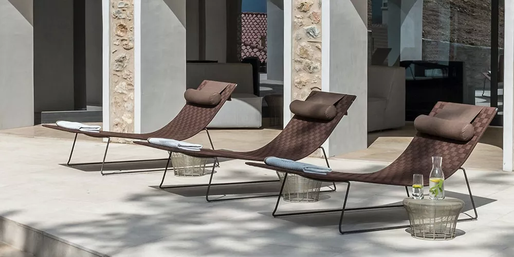 Drei braune Andreu World Tendra Sonnenliegen auf einer Terrasse mit Steinmauer im Hintergrund, jede mit einem Handtuch und Kissen.