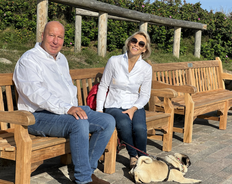 Zwei Personen in weißen Hemden sitzen auf einer Holzbank im Freien, mit einem Hund an der Leine auf dem Boden.