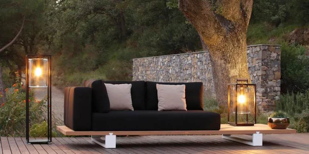 Gartenstimmung mit Villa Schmidt Gartenleuchten, schwarz, neben einem modernen Sofa auf einer Holzterrasse vor einer Steinmauer.