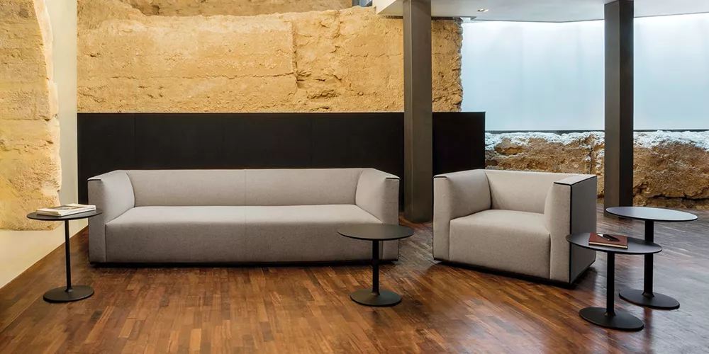 Andreu World Dual Sofa und Sessel in Grau auf Holzboden vor rustikaler Steinwand, begleitet von runden schwarzen Beistelltischen.