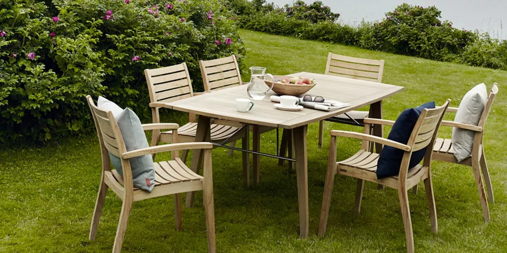 Sechs Skagerak Ballare Gartenstühle und ein Tisch aus hellem Holz auf einer grünen Wiese mit blühenden Büschen im Hintergrund.
