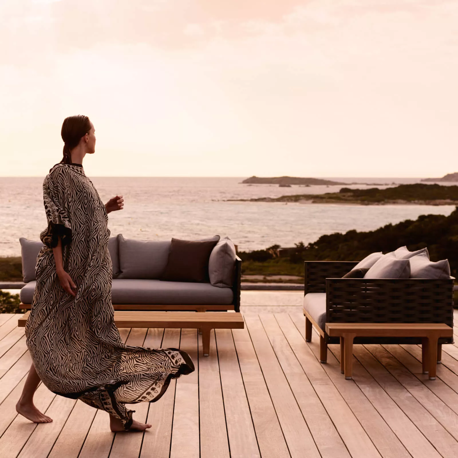 Eine Frau in einem langen Kleid geht an der Dedon Sealine Lounge vorbei auf einer Terrasse mit Meerblick im Hintergrund 