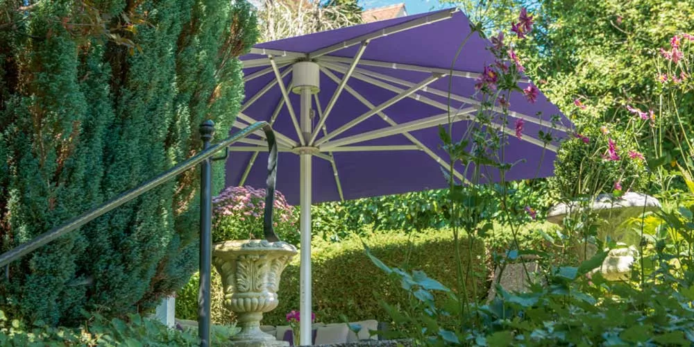 Glatz Palazzo Noblesse Sonnenschirm in Lila, im Garten aufgestellt, umgeben von grüner Vegetation und Blumen, bei sonnigem Wetter.