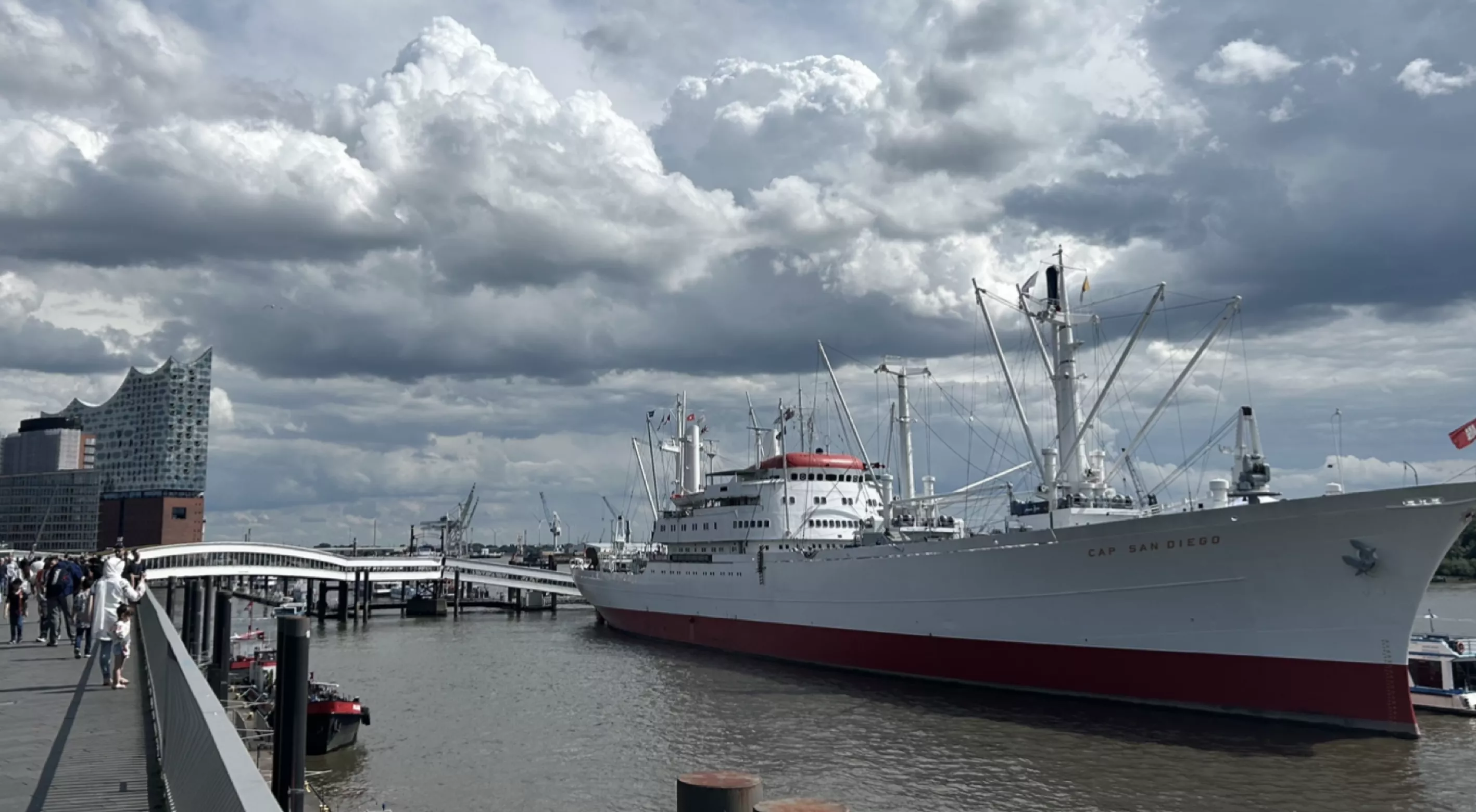 Ein weiß-rotes Schiff, die Cap San Diego, liegt im Hamburger Hafen vor Anker, mit der Elbphilharmonie im Hintergrund unter bewölktem Himmel.