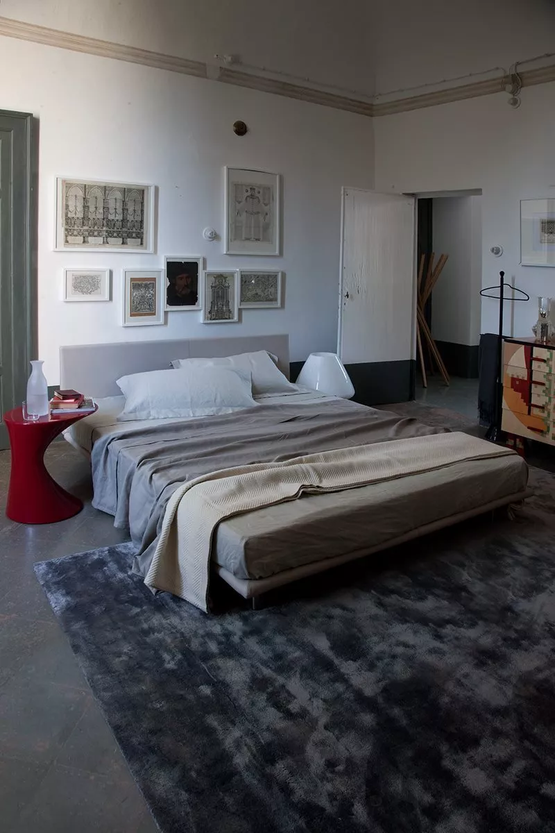 Ein gemütliches Schlafzimmer mit einem Zanotta Bett und einem roten Beistelltisch auf einem grauen Teppich mit Kunstwerken an der Wand.