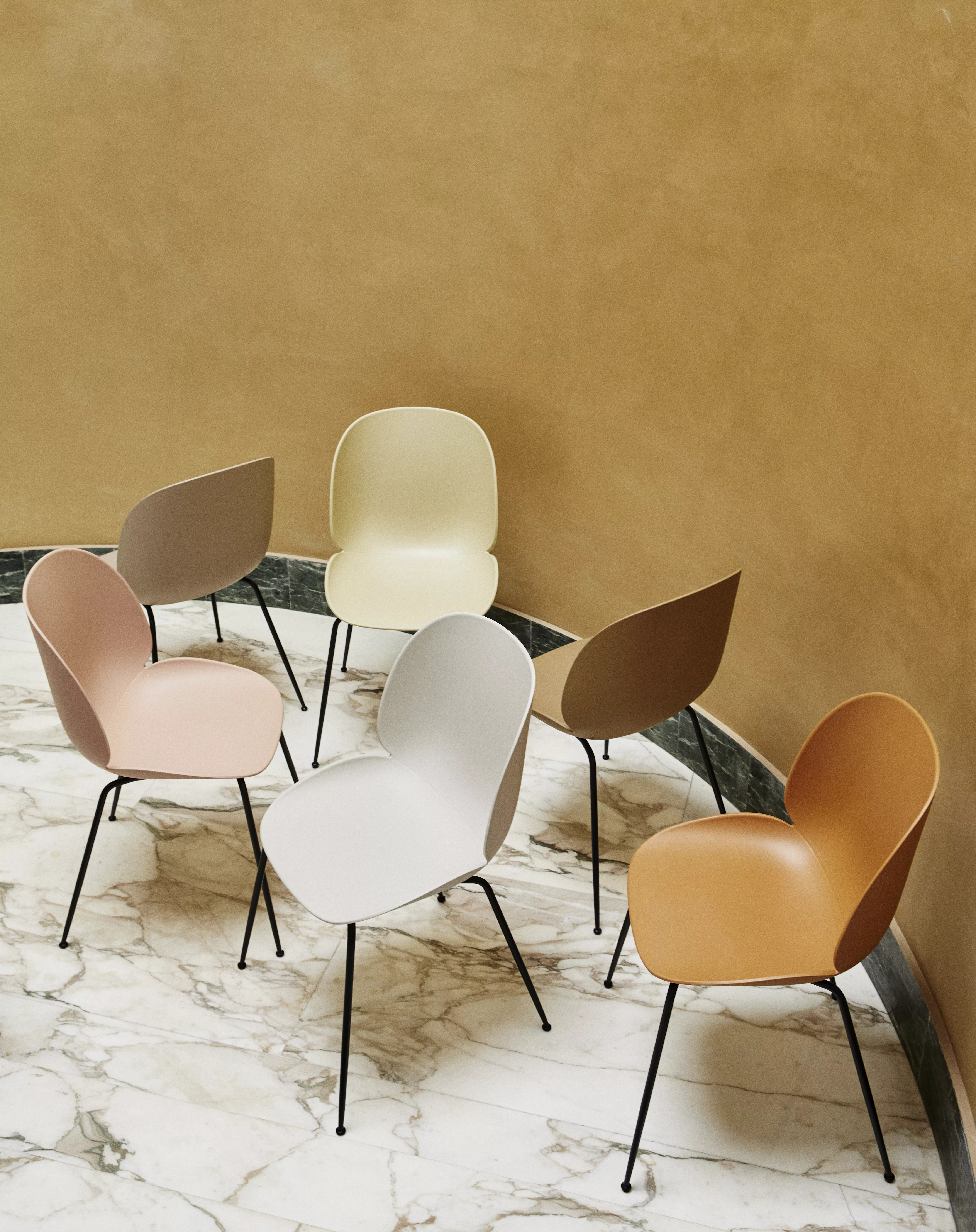 Sechs Gubi Stühle in verschiedenen Farben stehen auf einem Marmorboden in einem Raum mit beiger Wand in einer kreisförmigen Anordnung