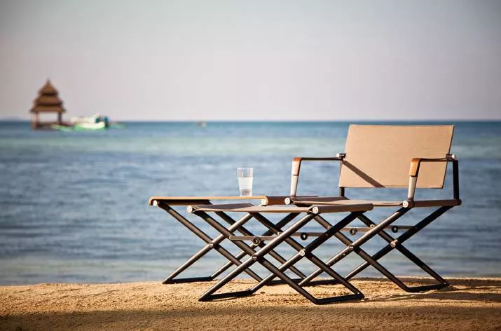 Dedon Seax Armlehnstuhl und Tisch am Strand mit einem Glas Wasser auf dem Tisch im Hintergrund das Meer und ein Boot mood picture