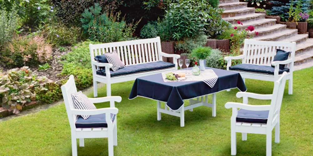 Weiße Aerrenhaus Antoine Essgruppe im Garten, bestehend aus Tisch, zwei Bänken und zwei Stühlen mit schwarzen Kissen, auf grünem Rasen.