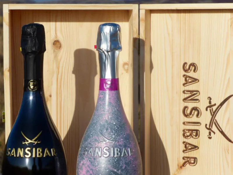Sansibar Sylt Prosecco 2er-Set in Holzkiste, eine Flasche schwarz, eine silber mit Glitzer, vor hellem Hintergrund.