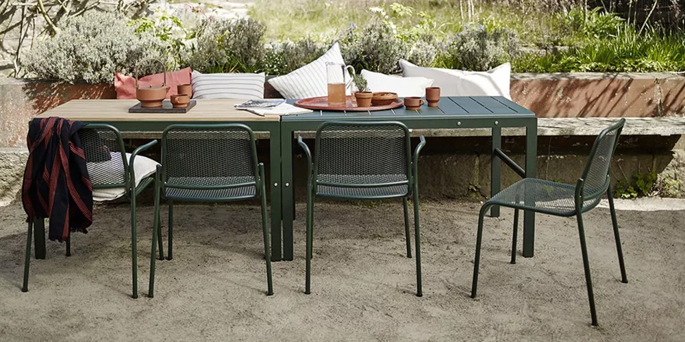 Skagerak Mira Gartenmöbel-Set in Dunkelgrün auf Sandboden, umgeben von Kissen und Pflanzen im Hintergrund, stimmungsvolle Outdoor-Szene.