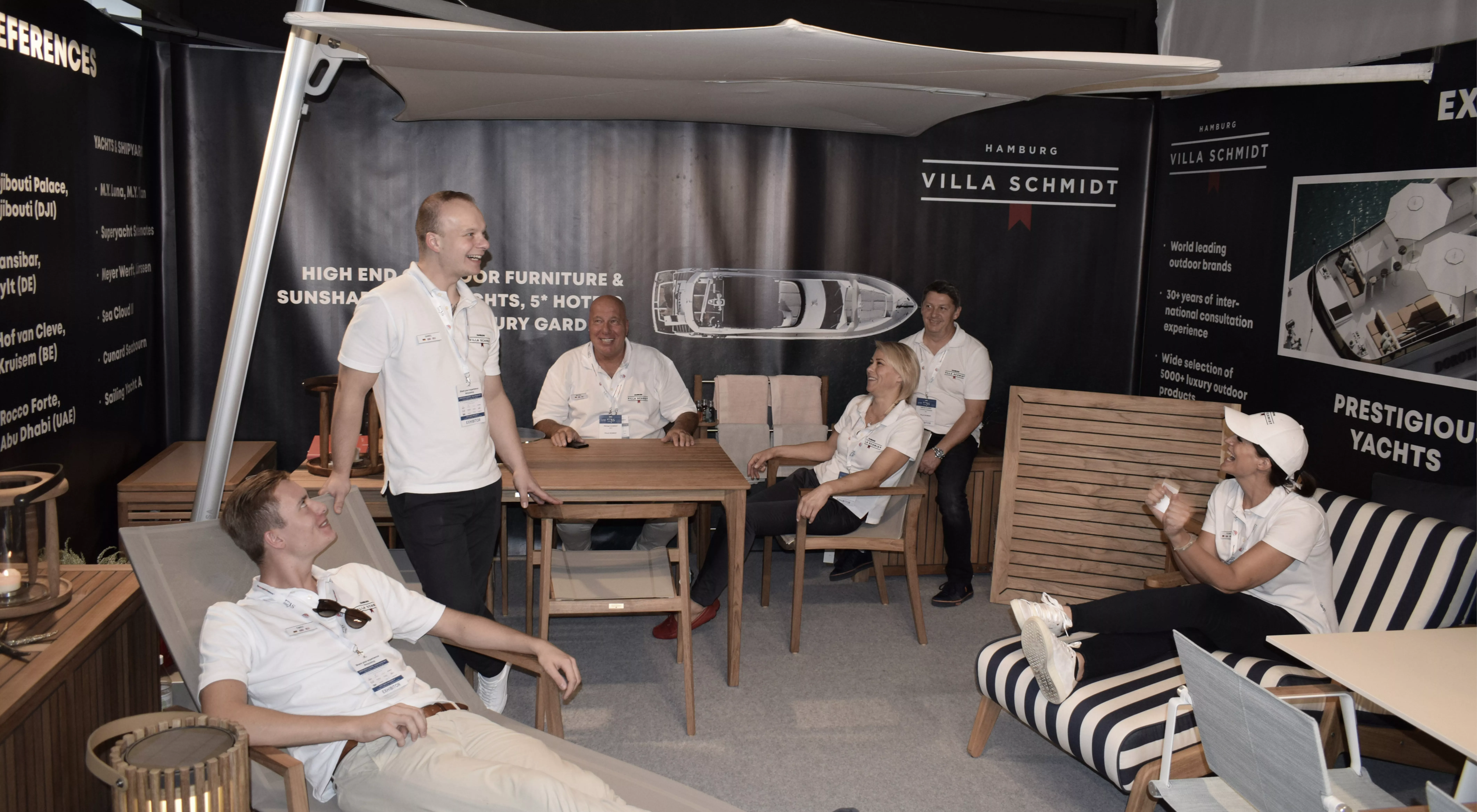 Personen in einem Ausstellungsraum von Villa Schmidt mit Gartenmöbeln und Sonnenschirmen, umgeben von Werbebannern und Prospekten.