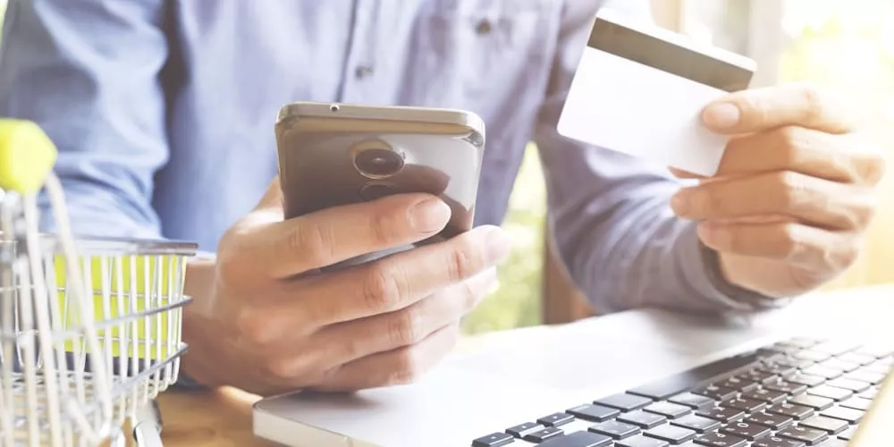 Person in blauem Hemd hält Smartphone und Kreditkarte vor Laptop, Einkaufswagen im Vordergrund, helle Hintergrundbeleuchtung.