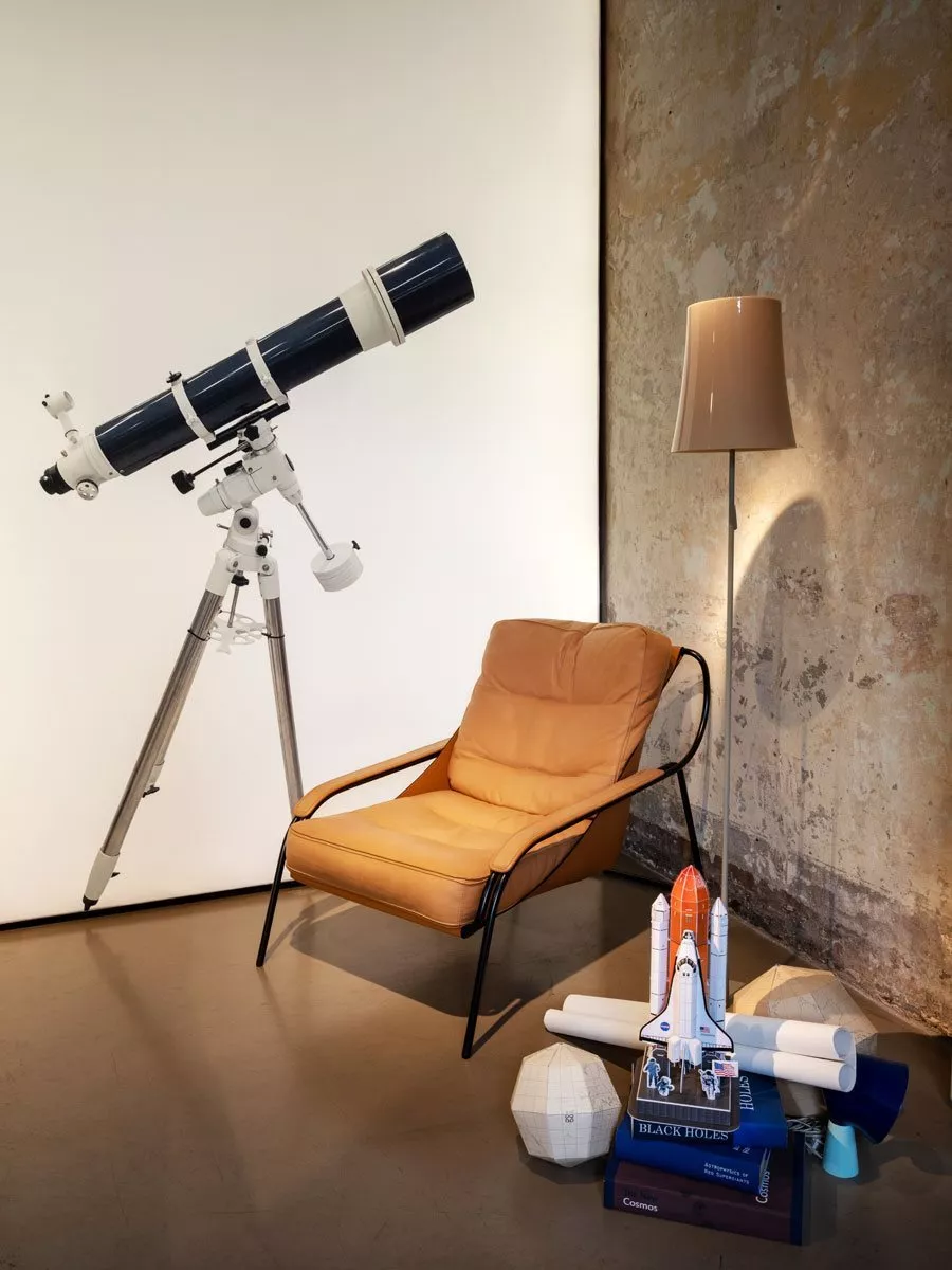 Ein gemütlicher Raum mit einem Zanotta 900 Maggiolina Sessel einer Stehlampe einem Teleskop und einem Modell eines Space Shuttles auf Büchern.