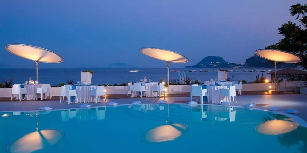 Stimmungsbild mit Eclipsum Sonnenschirmen in Weiß bei Sonnenuntergang, auf einer Terrasse mit Blick auf das Meer und gedeckten Tischen.