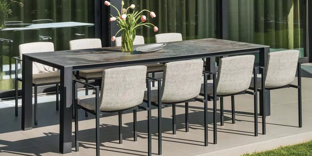 Gartenszene mit Calma Ten Esstisch in Schwarz und acht beigefarbenen Stühlen auf einer Terrasse vor einer Glasfassade.