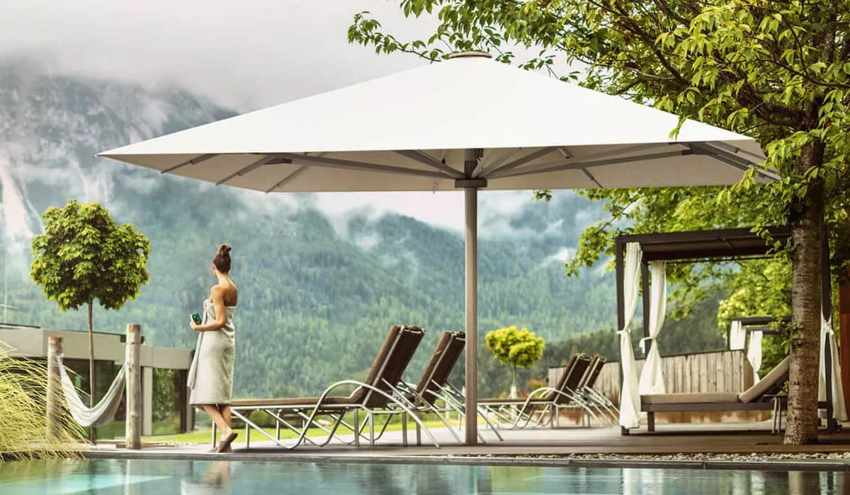 Frau in Garten mit Glatz Sonnenschirm Palazzo Noblesse in Weiß, umgeben von Liegestühlen und Bäumen, vor Bergkulisse.