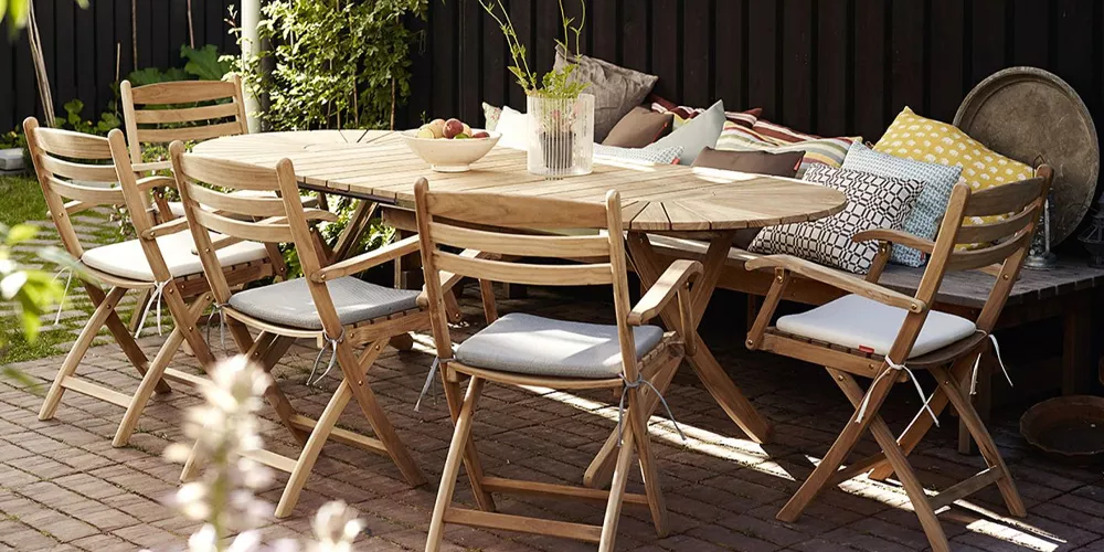 Skagerak Selandia Gartenmöbel-Set aus Teakholz auf einer Terrasse mit Kissen und Pflanzen im Hintergrund, stimmungsvolle Außenaufnahme.