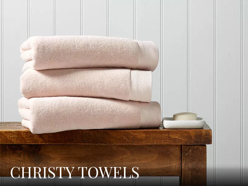 Christy Towels Prämie in zartem Rosa, gestapelt auf einem rustikalen Holztisch vor einer weißen Holzvertäfelung.