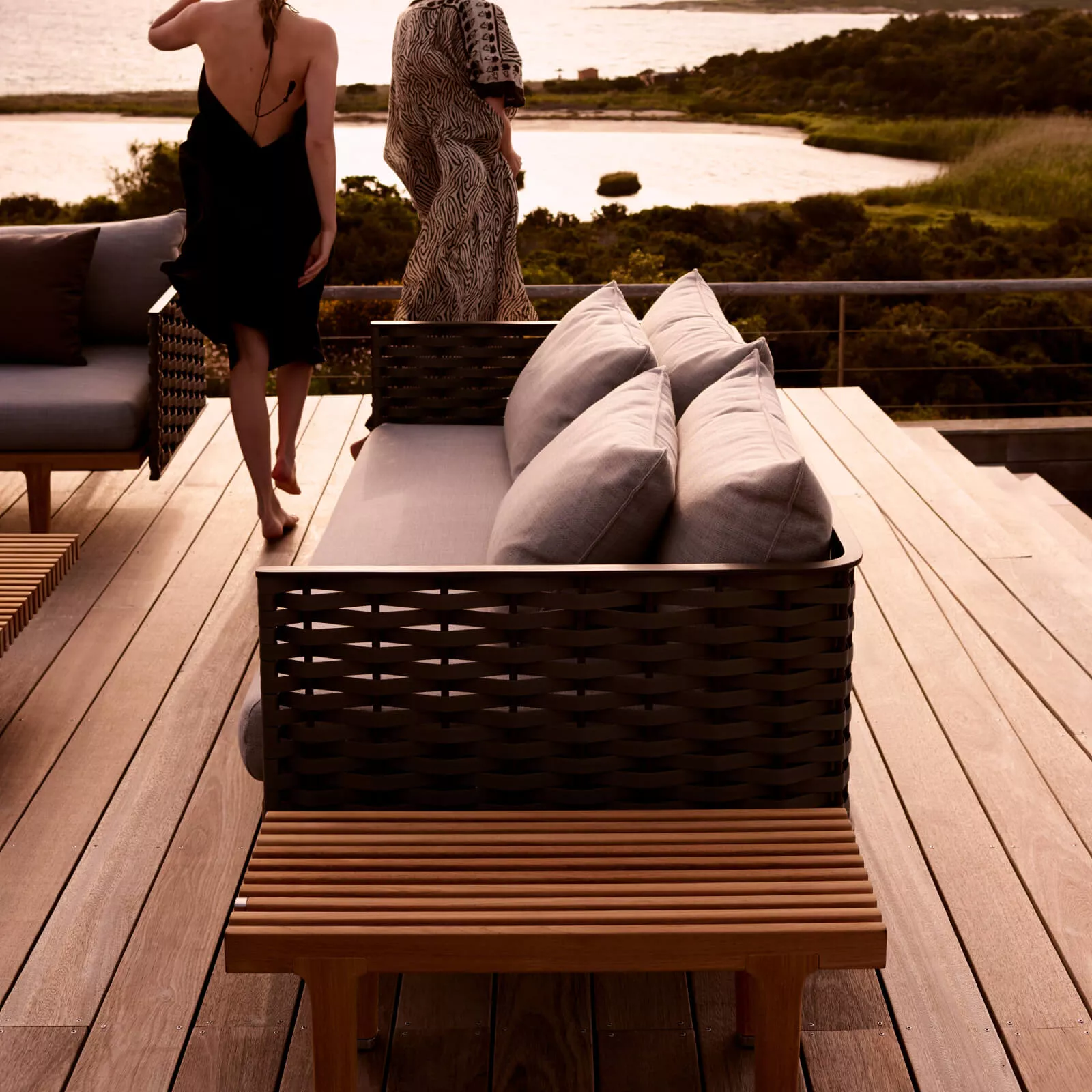 Zwei Frauen in Sommerkleidern gehen auf einer Terrasse mit Blick auf das Meer neben einem Dedon Sealine Loungesofa mit grauen Kissen.
