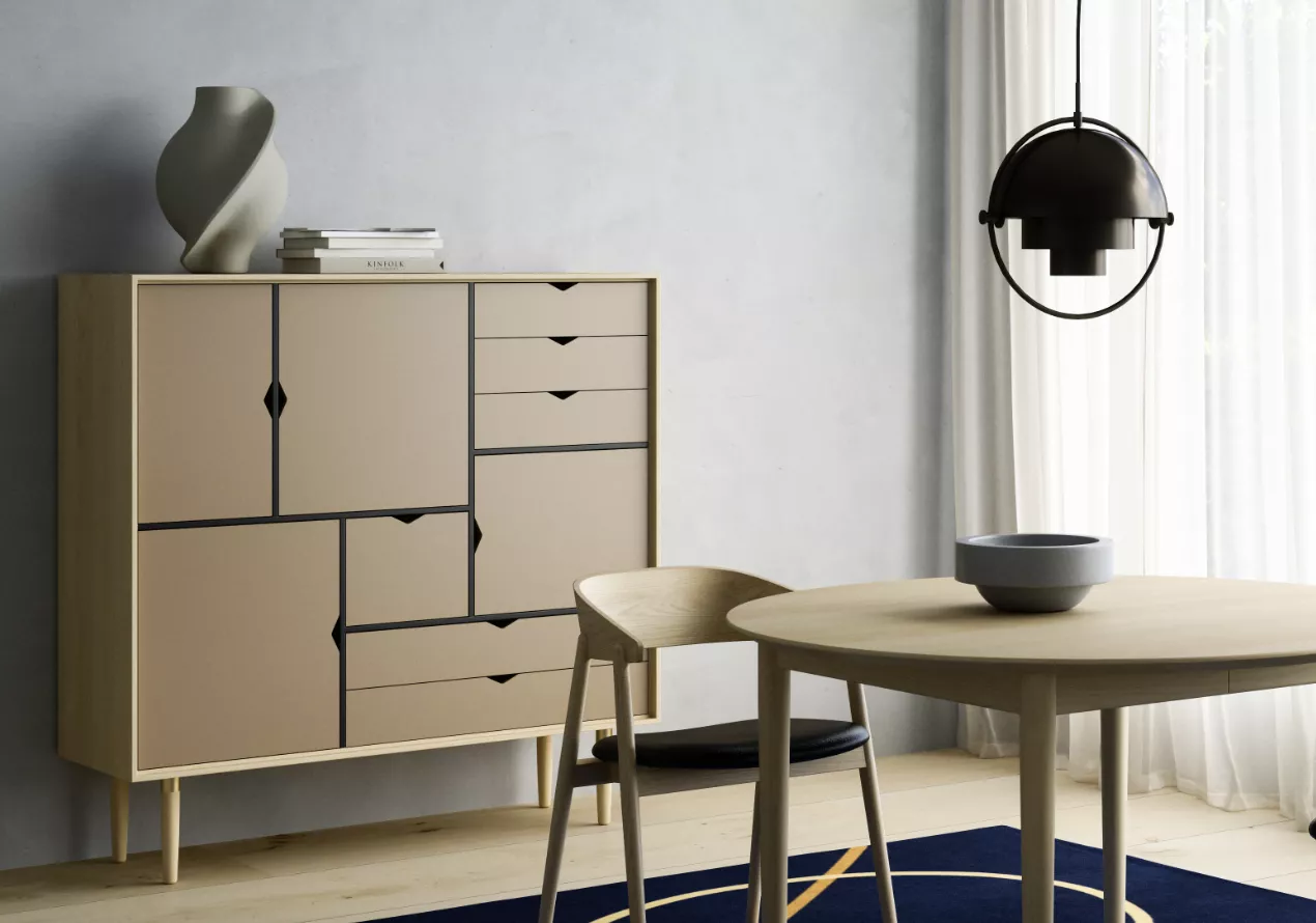 Beige "S Kollektion" Schrank von Villa Schmidt in modernem Wohnzimmer mit hellem Holztisch und Stuhl, vor grauer Wand und Vorhängen.